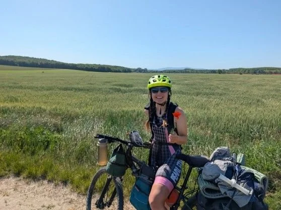 Skizzi in a green helmet standing with her loaded bike on a dirt track beside wide green fields under a clear sky