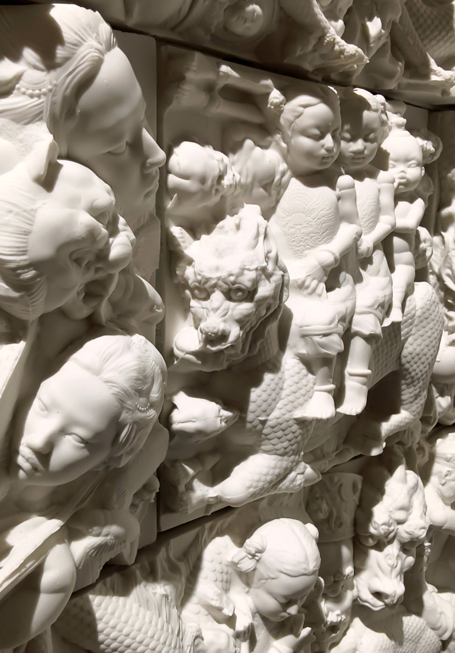White ceramic relief with clustered human faces, figures and animal forms.