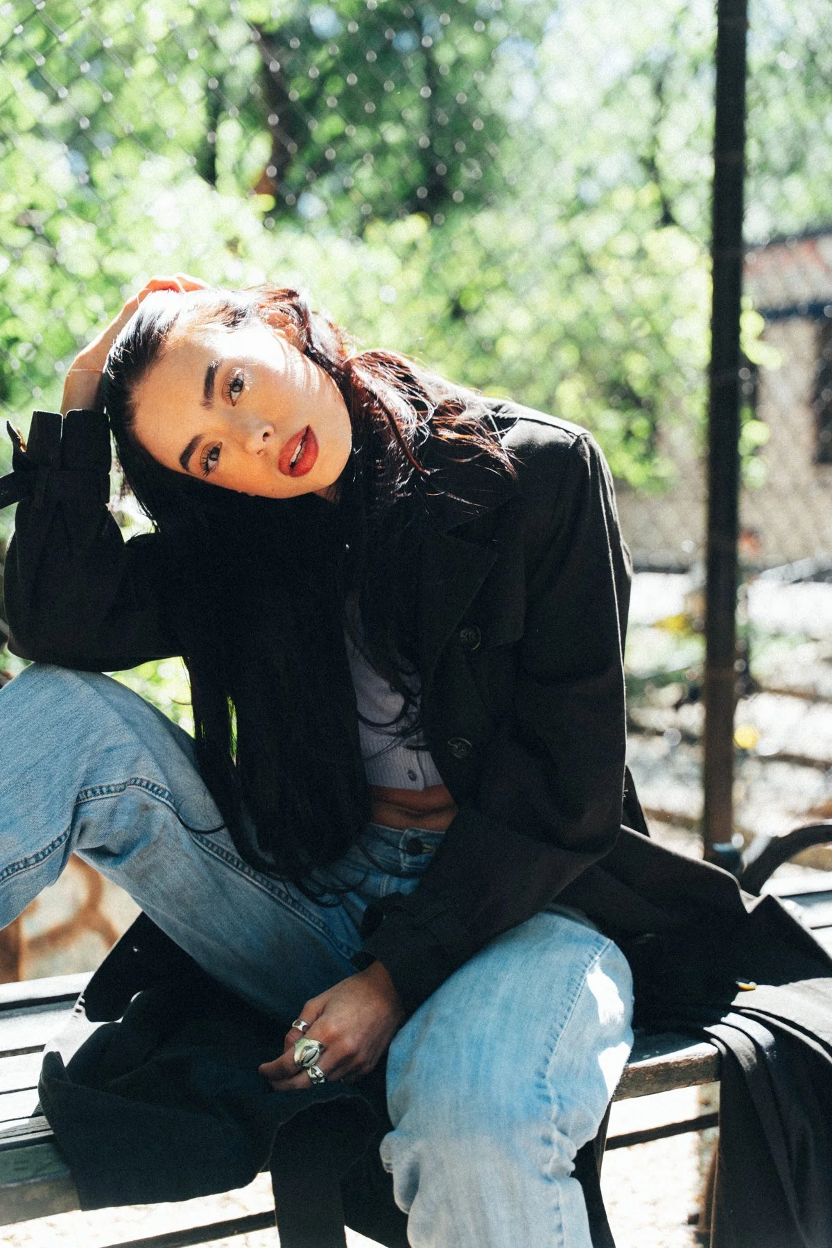 Hannahbeth seated on a bench in a black jacket and jeans, with sunlight on her face and a leafy fence behind her