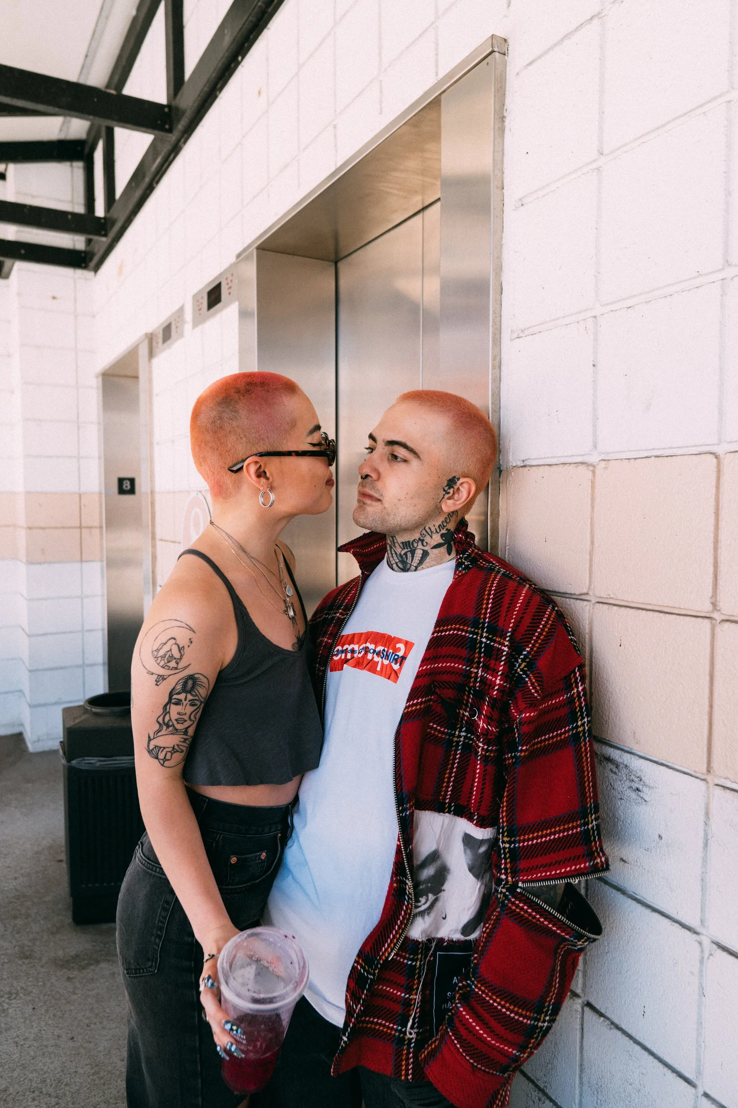 Ash and AJ leaning close together beside a white brick wall