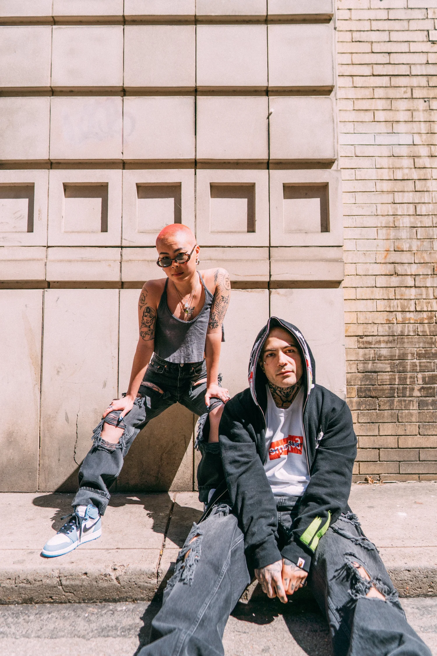 Ash and AJ seated against a pale masonry wall in layered casual clothing