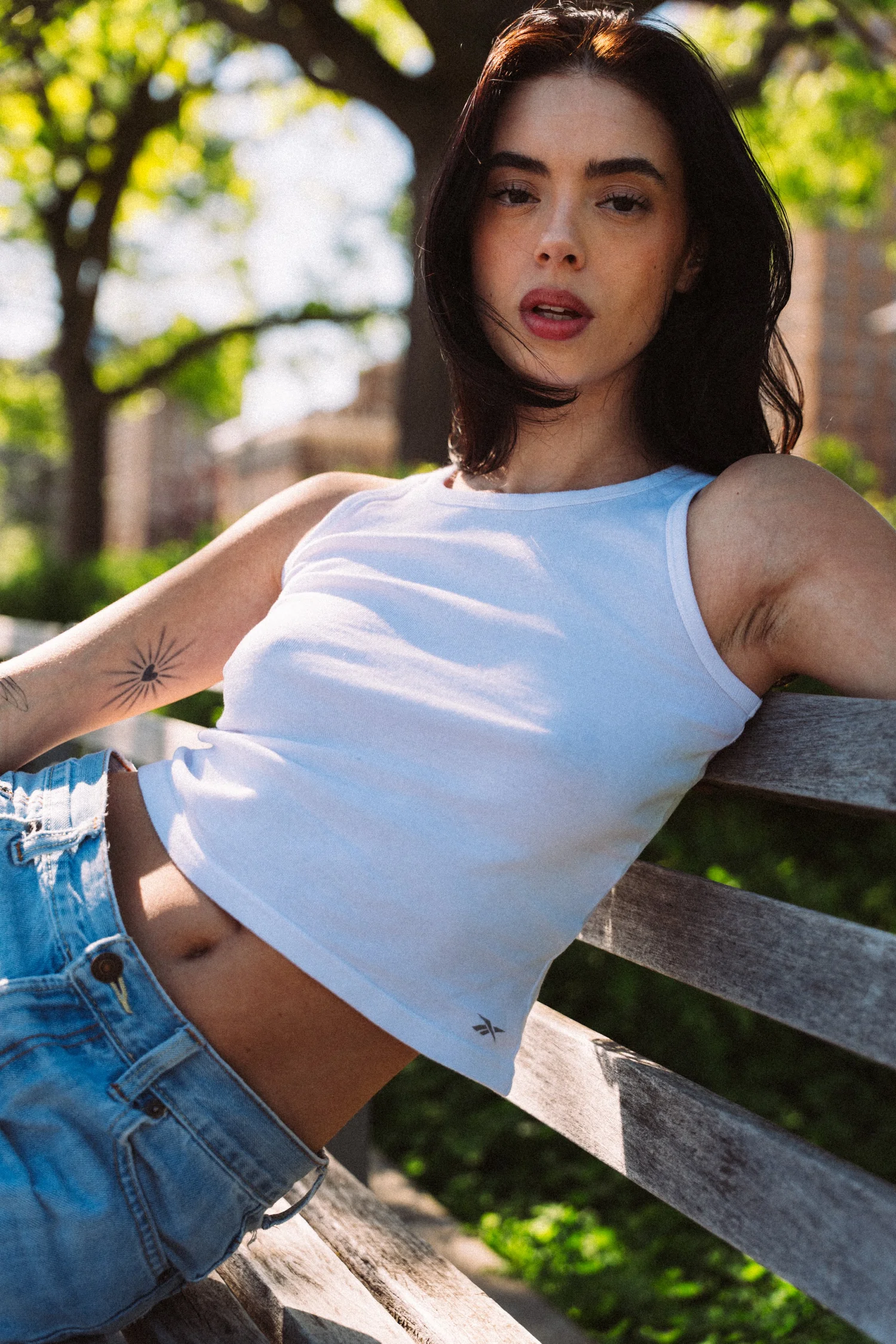 Hannahbeth leaning along a wooden bench beneath trees, with sunlight catching her hair