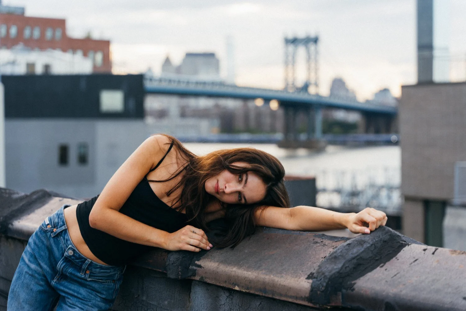 Amanda leaning sideways across a rooftop ledge with a bridge behind her