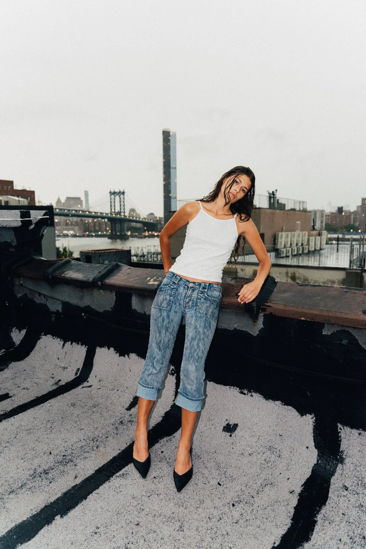 Full-body portrait of Tara in a white tank top, cuffed jeans, and black heels on a rooftop with the skyline behind her