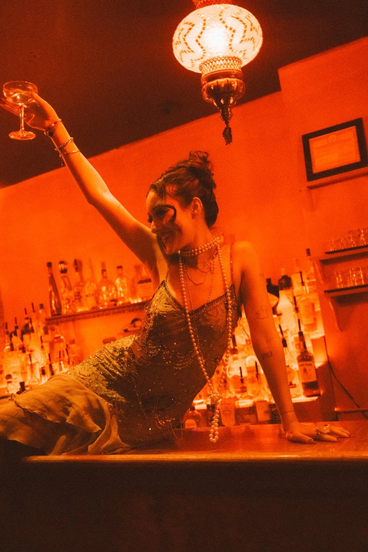 Eden seated on the bar with an arm raised beneath a hanging lantern