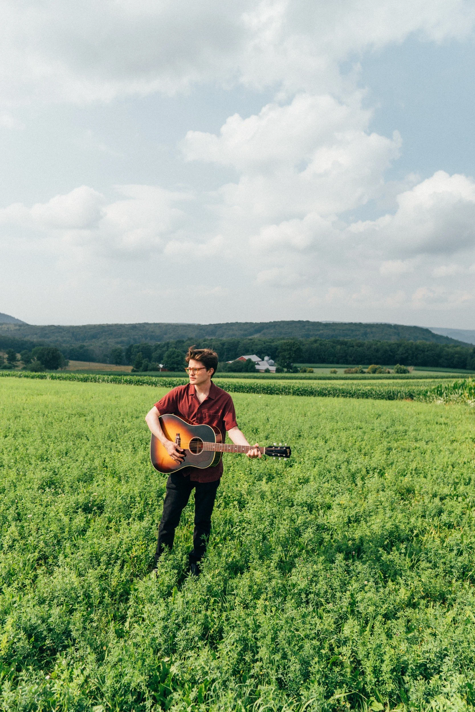Zack Keim holding an acoustic guitar in a green field beneath a cloudy sky.