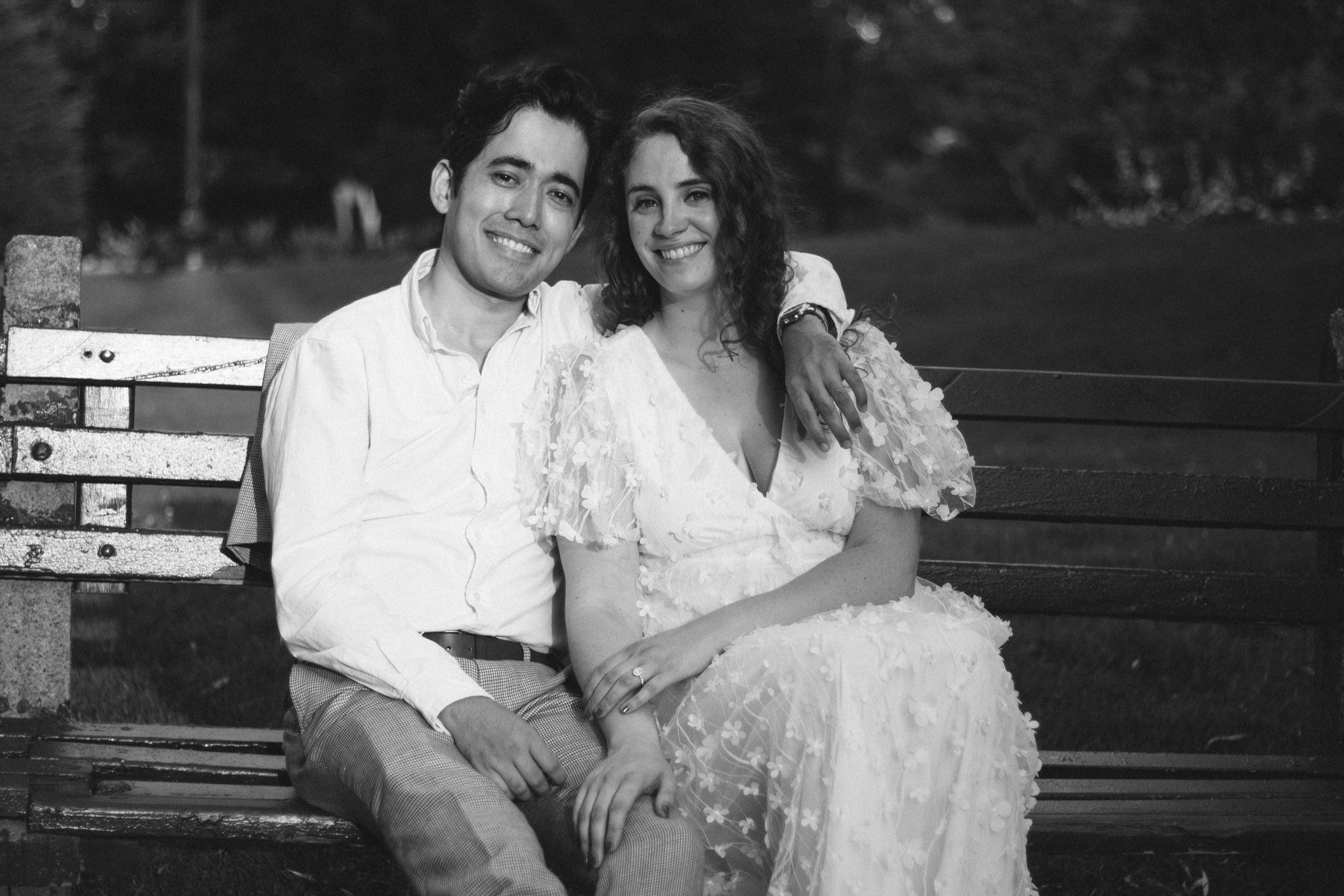 Black-and-white portrait of Stefano and Maddy smiling together on a park bench
