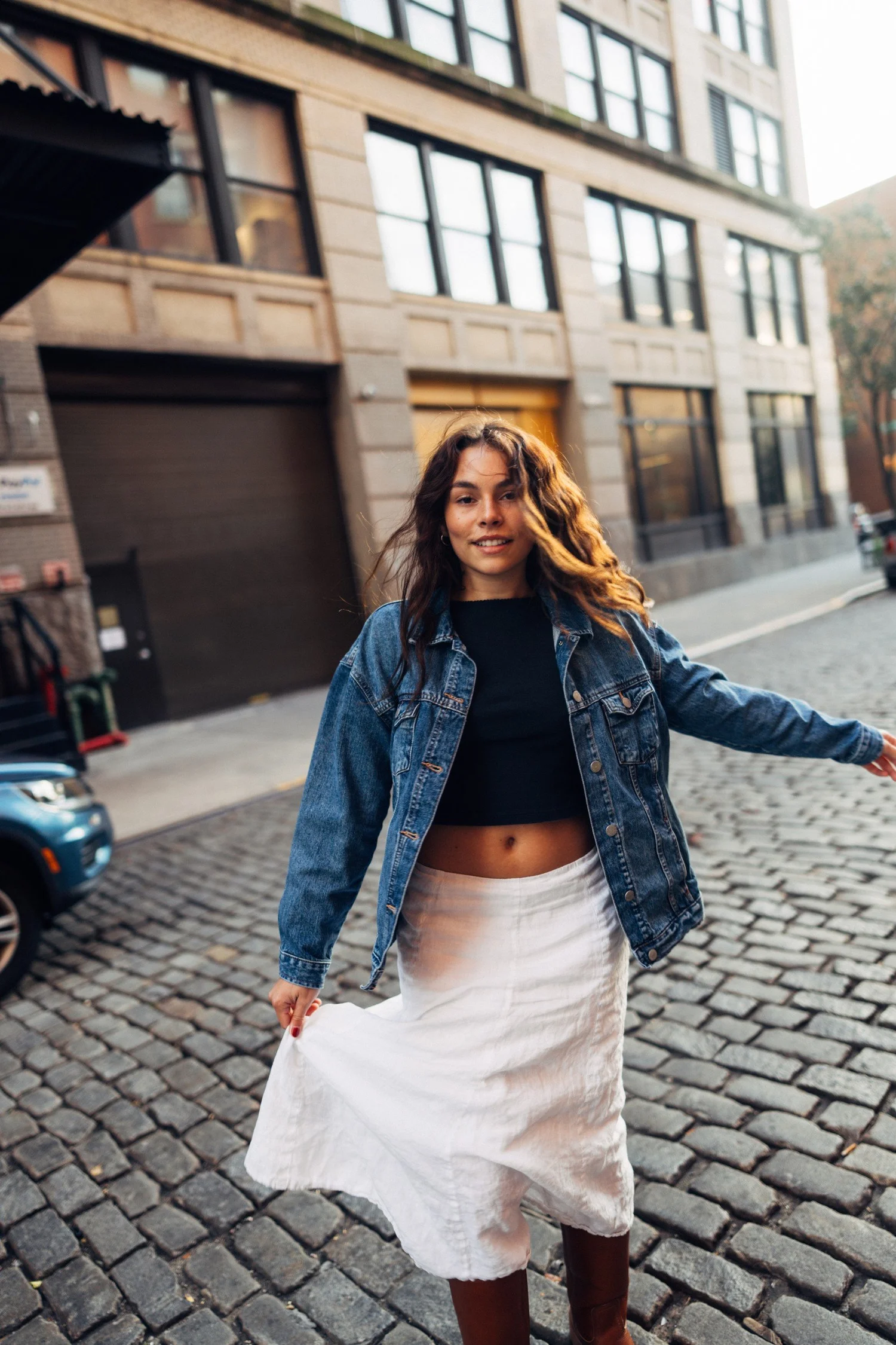 Marley in a denim jacket and white skirt with one arm extended on a cobbled street.