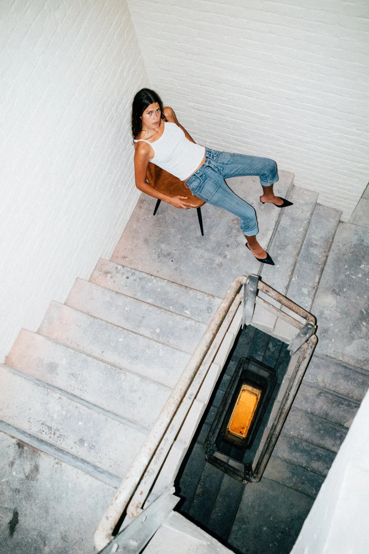 Tara sitting in a brown chair beside a stairwell in a white brick interior