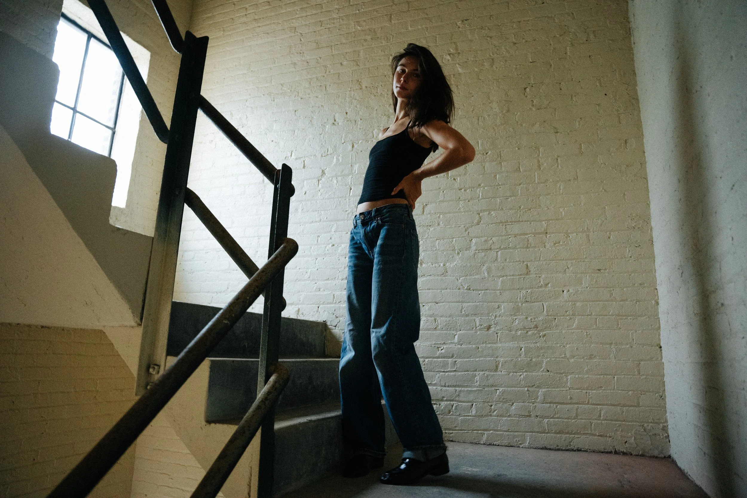 Amanda standing beside a stair railing and window in a white brick interior.