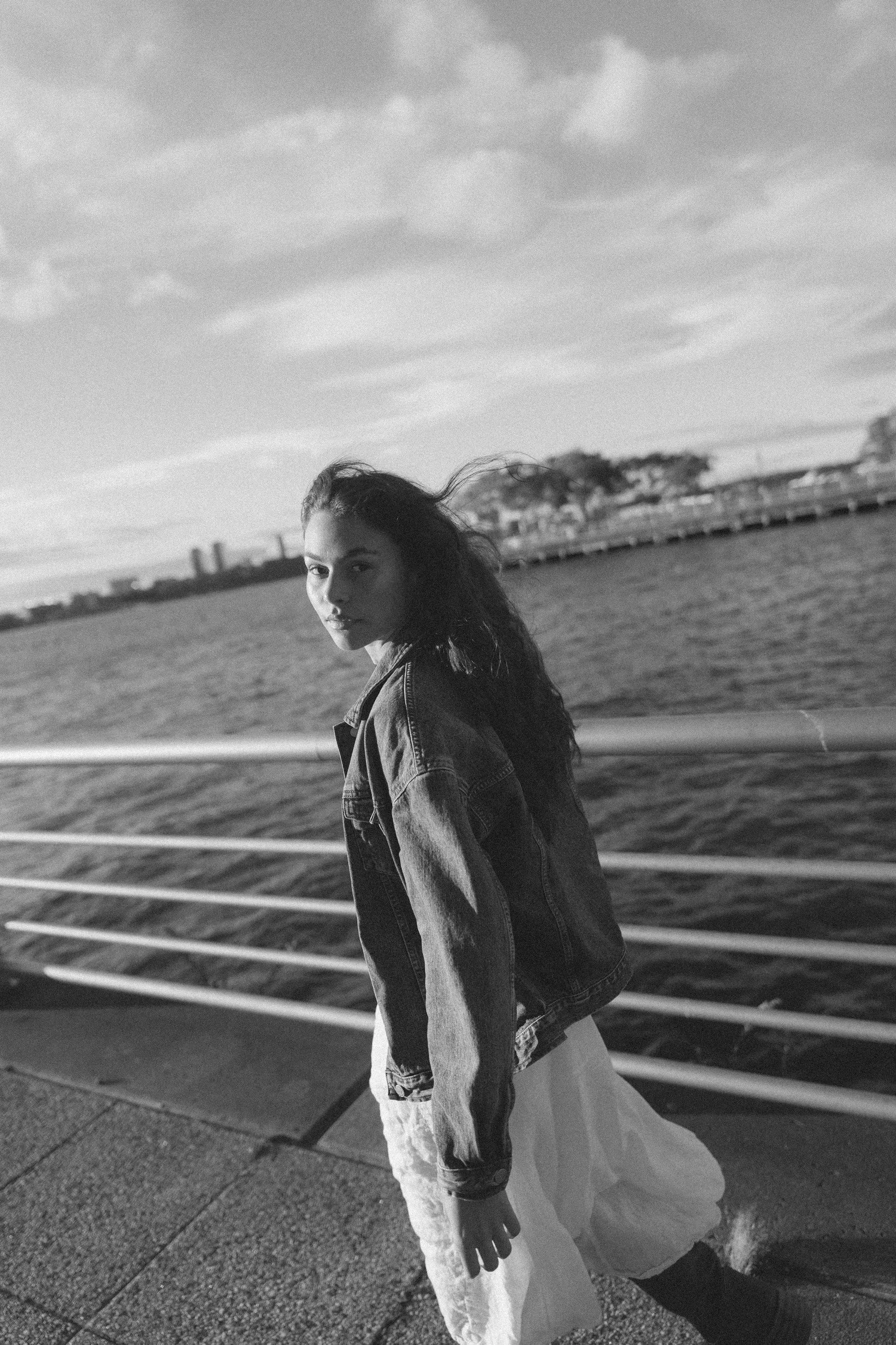 Black-and-white portrait of Marley in a denim jacket and pale skirt beside a waterfront railing.