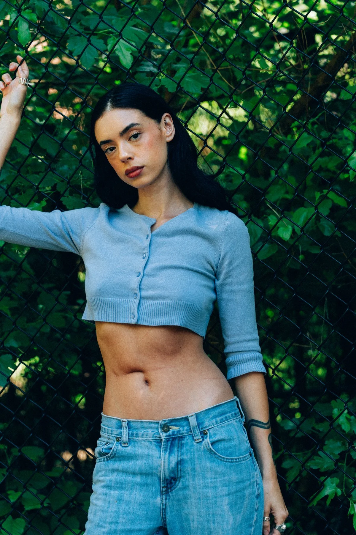 Hannahbeth in a light blue cardigan with one arm raised against a green chain-link fence
