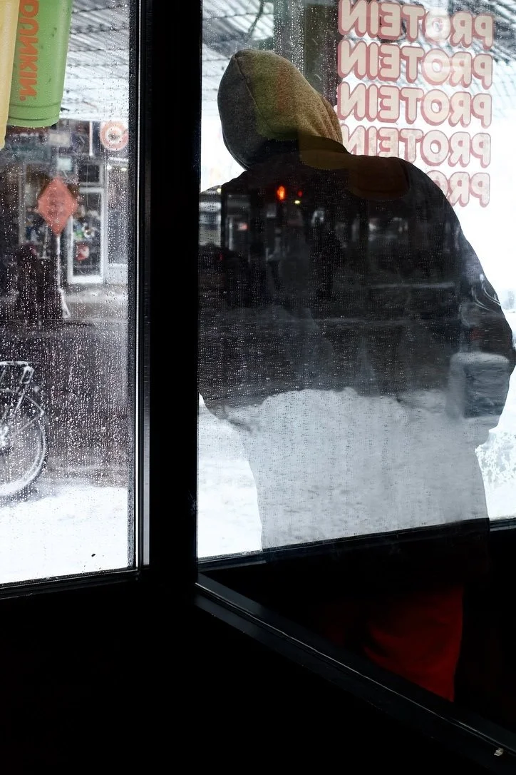 Hooded person seen through a wet Dunkin window layered with reflections