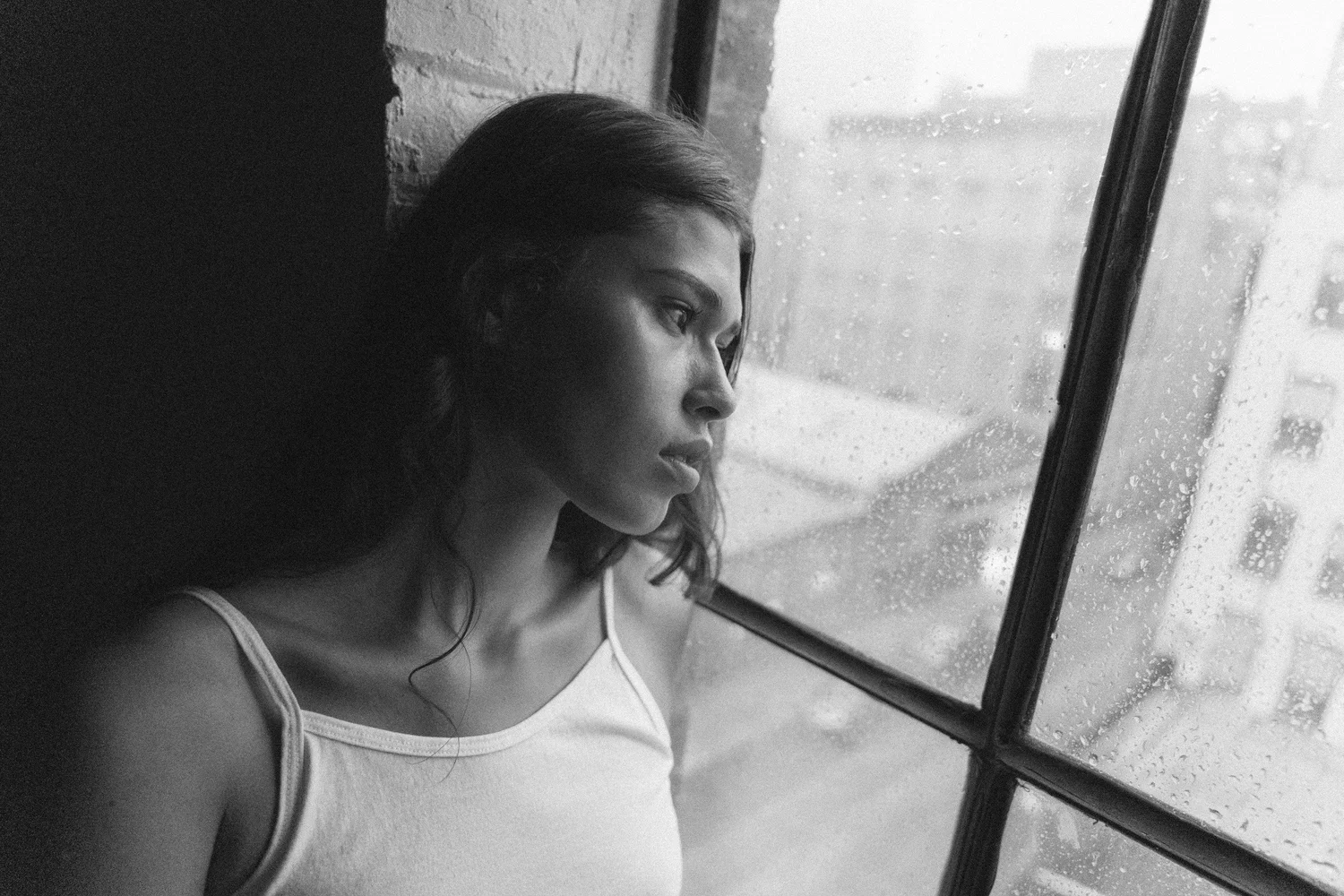 Black-and-white portrait of Tara looking through a window in a white tank top.