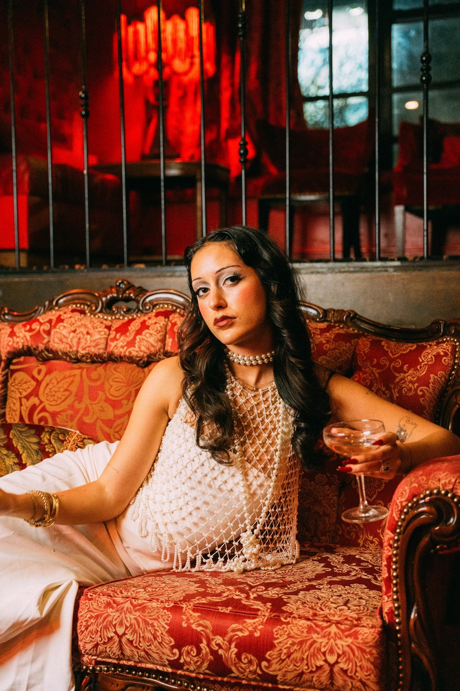 Eden seated in an ivory dress on a patterned red sofa beneath lanterns