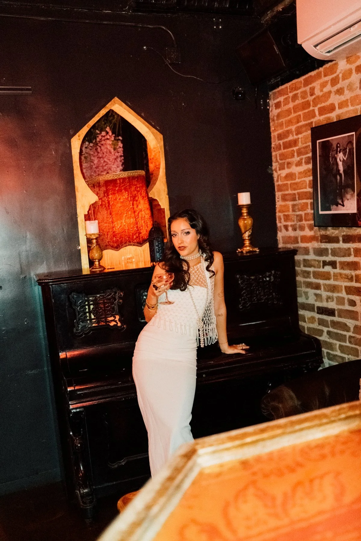Eden in a white dress leaning against a dark upright piano beneath an ornate arch