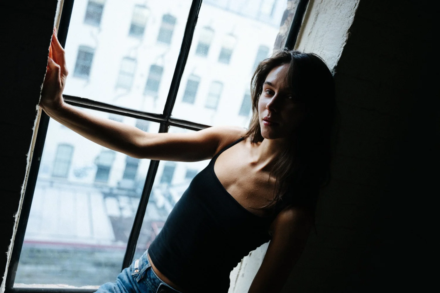 Amanda in a black tank top beside a bright window, photographed at a diagonal angle in a dark interior