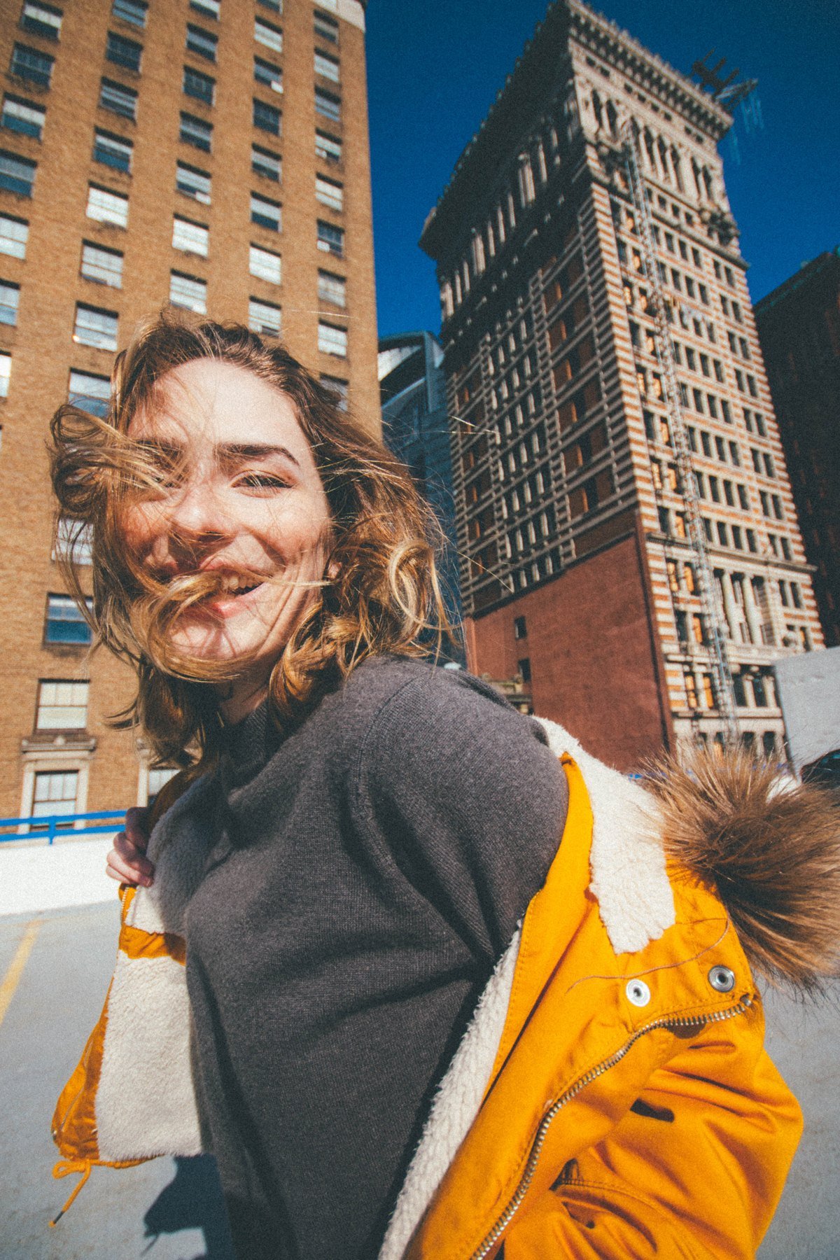 Sarah with windblown hair wearing a yellow coat and gray sweater against tall buildings