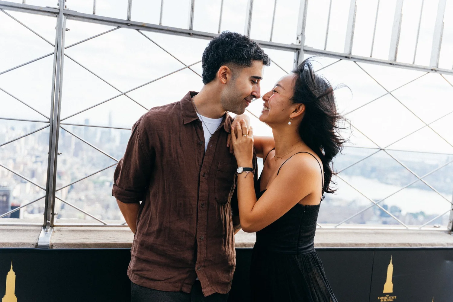 A smiling couple leaning close together at an outdoor viewing deck, with city buildings beyond the fence