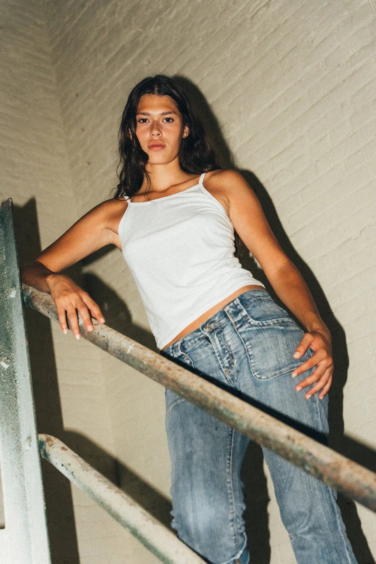 Tara leaning beside a stair railing in a white tank top and jeans