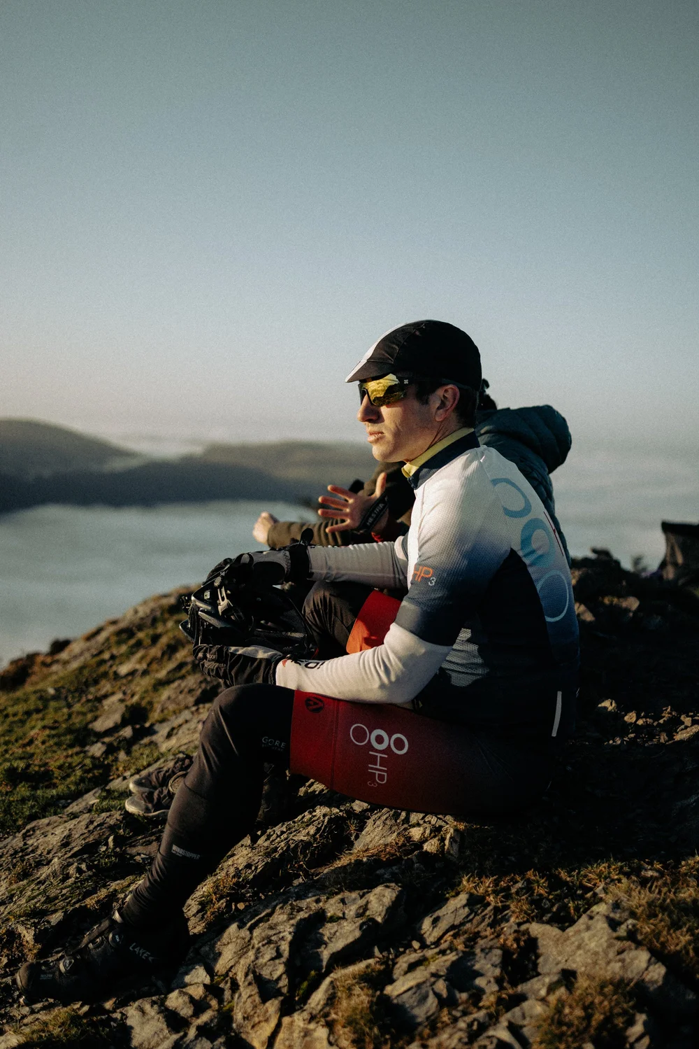 An HP3 athlete sitting on a summit at sunrise, looking out over a cloud inversion