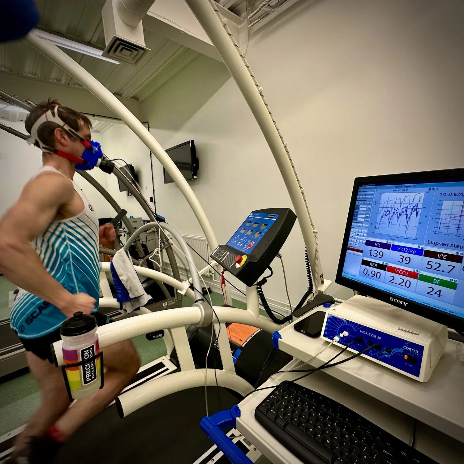 An athlete running on a treadmill in the exercise physiology lab, wearing a gas-analysis mask, with live oxygen and heart-rate data on the screen beside them
