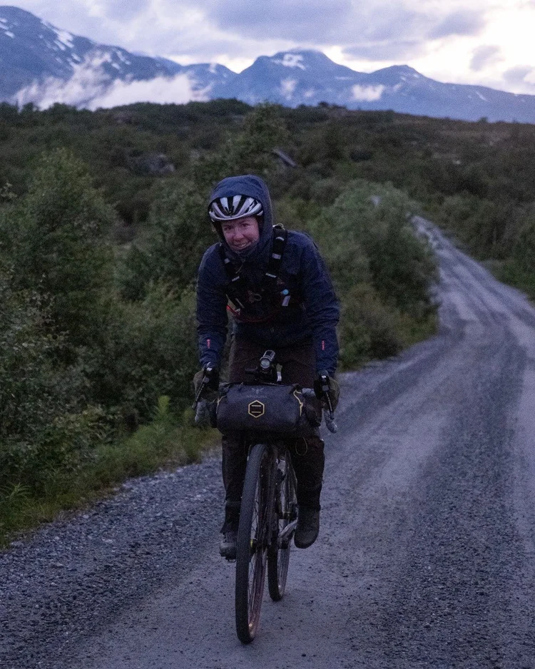 Clare Walkeden smiling as she rides a gravel road at dusk with mountains behind