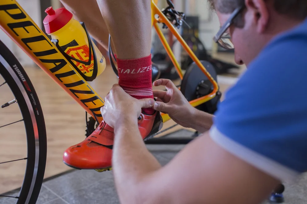 A physiotherapist checking the cleat position on a cyclist's shoe while they sit on the bike