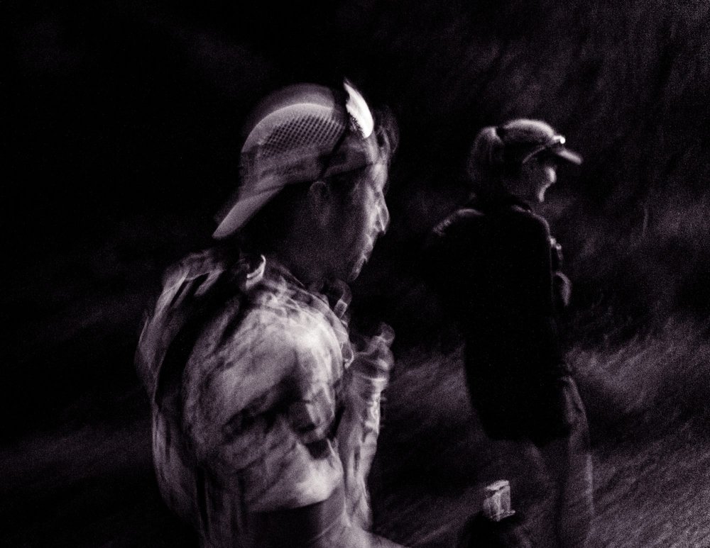 Two trail runners talking in the dark at a checkpoint, lit by head torch