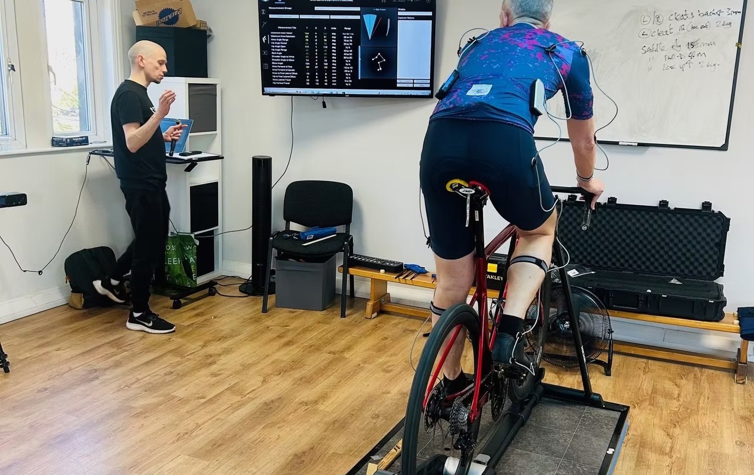 Will Beauchamp reviewing a rider's Retul 3D data on the laptop during a bike fit at Worsley Physiotherapy Clinic, with the measurements on the screen behind