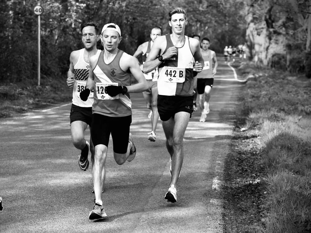 Three runners racing side by side on a tree-lined road, in black and white