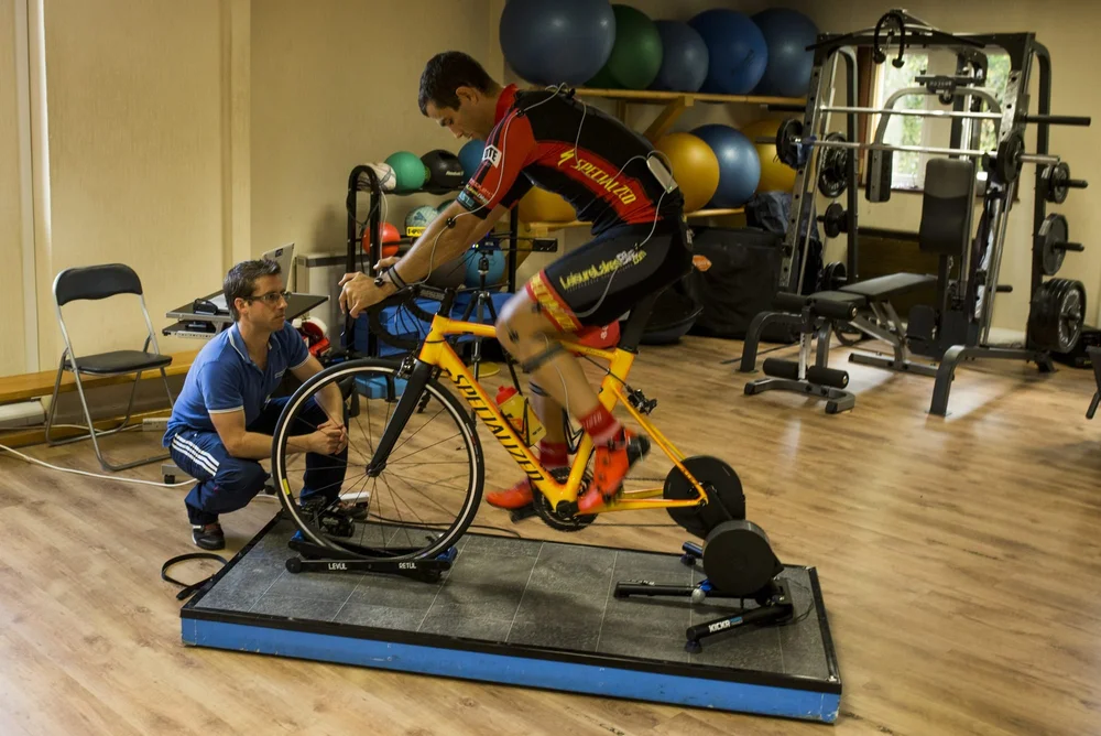 A cyclist riding their own bike on a turbo trainer in the clinic gym while the fitter watches from the side