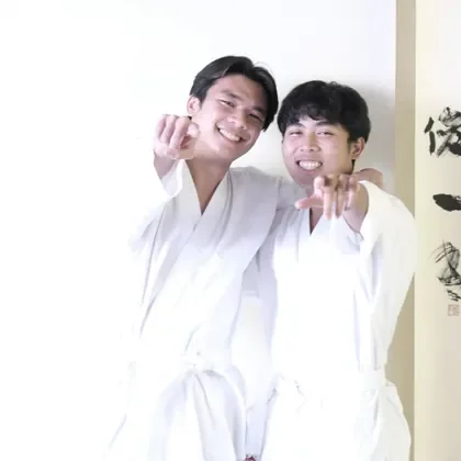Two Budo students in karate gi, arms around each other, pointing at the camera