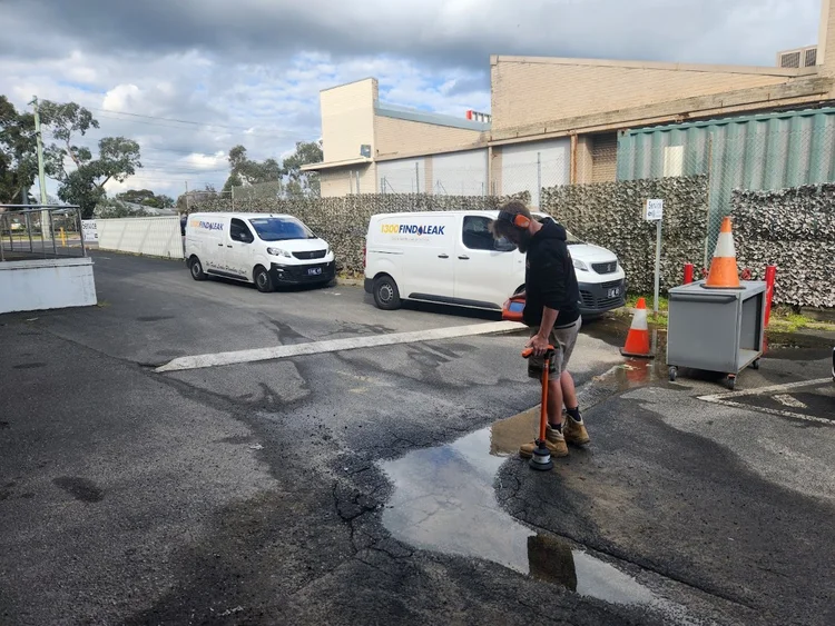 Technician listening for a burst water main on a wet commercial car park in Melbourne
