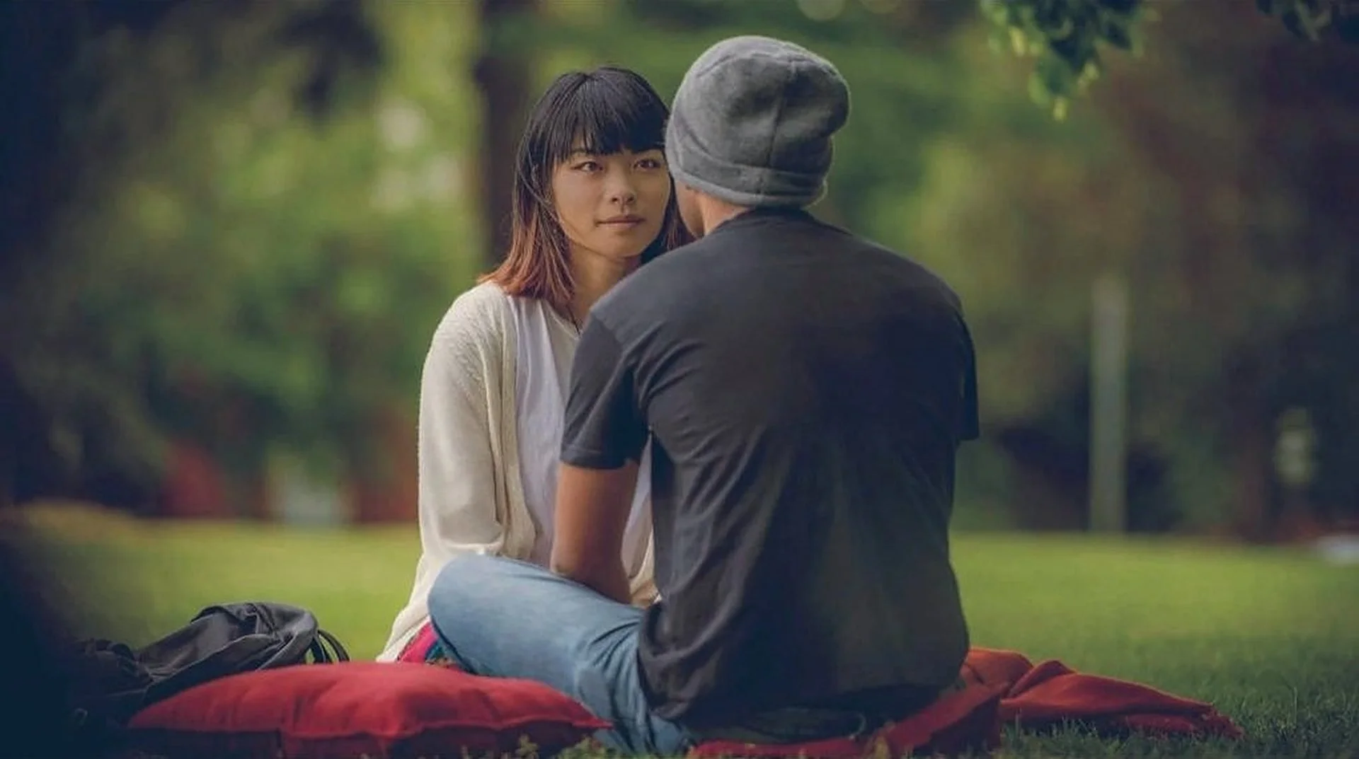 Two people on a red blanket in a park, holding each other's gaze