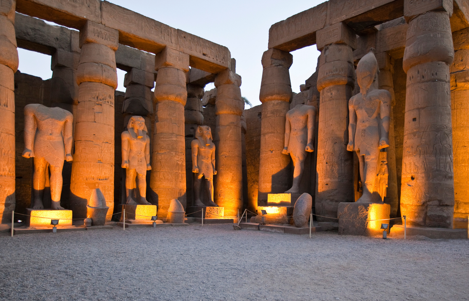 Floodlit colossal pharaoh statues between columns in a temple courtyard in Luxor at dusk