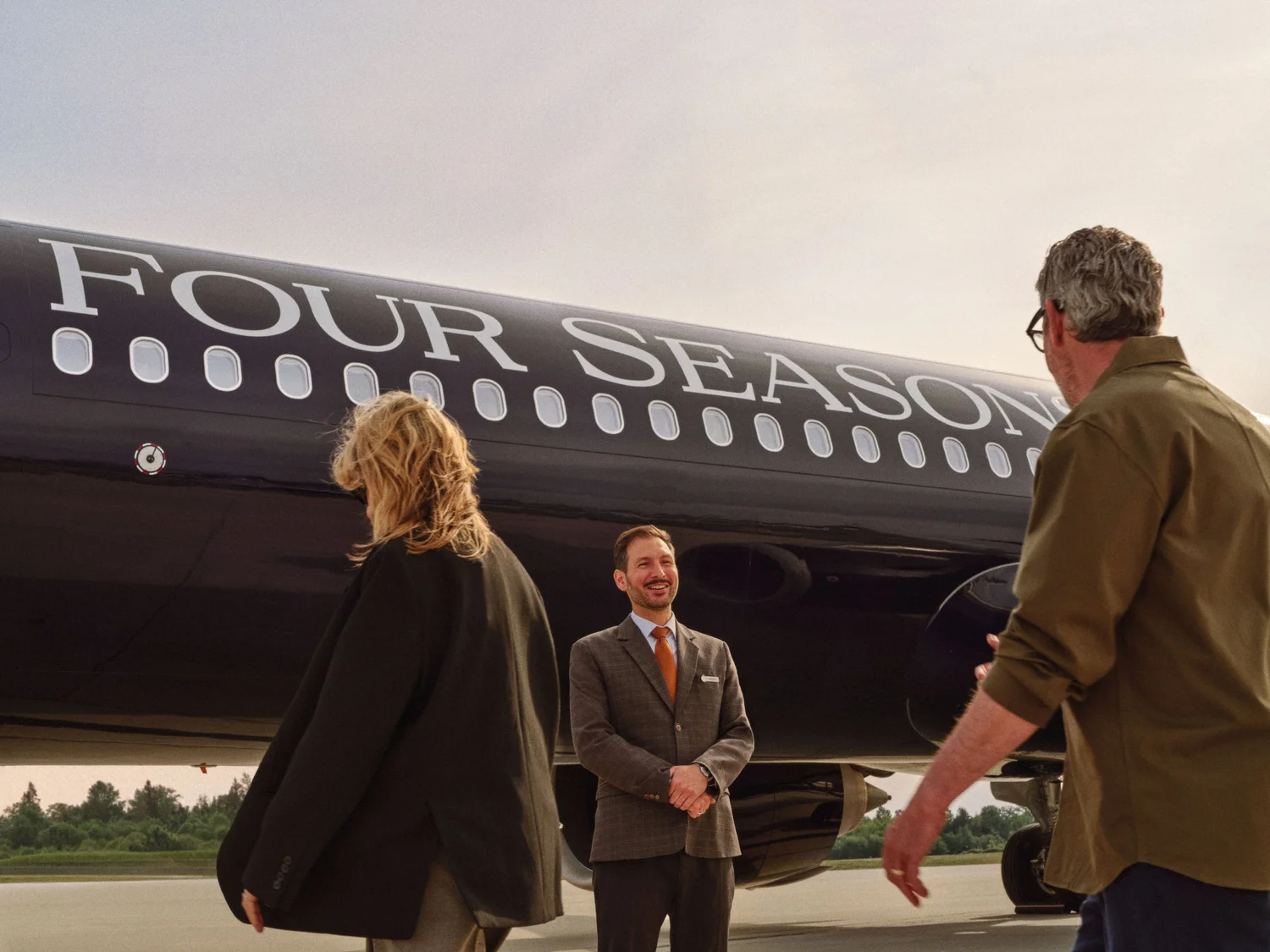 A Four Seasons host greets two guests on the tarmac beside the black Four Seasons Private Jet