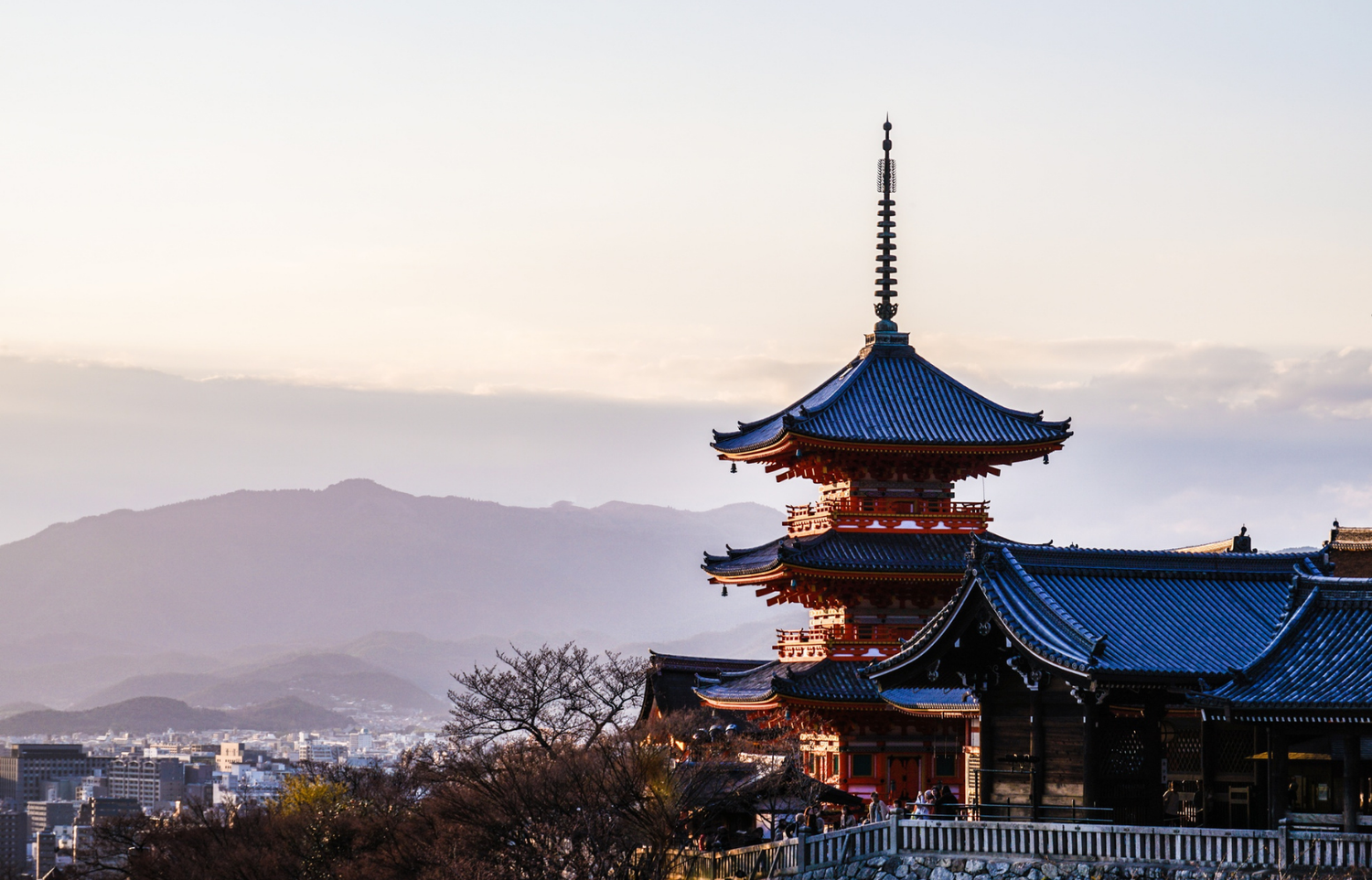 A red three-story pagoda above the rooftops of Kyoto at sunset, with mountains beyond