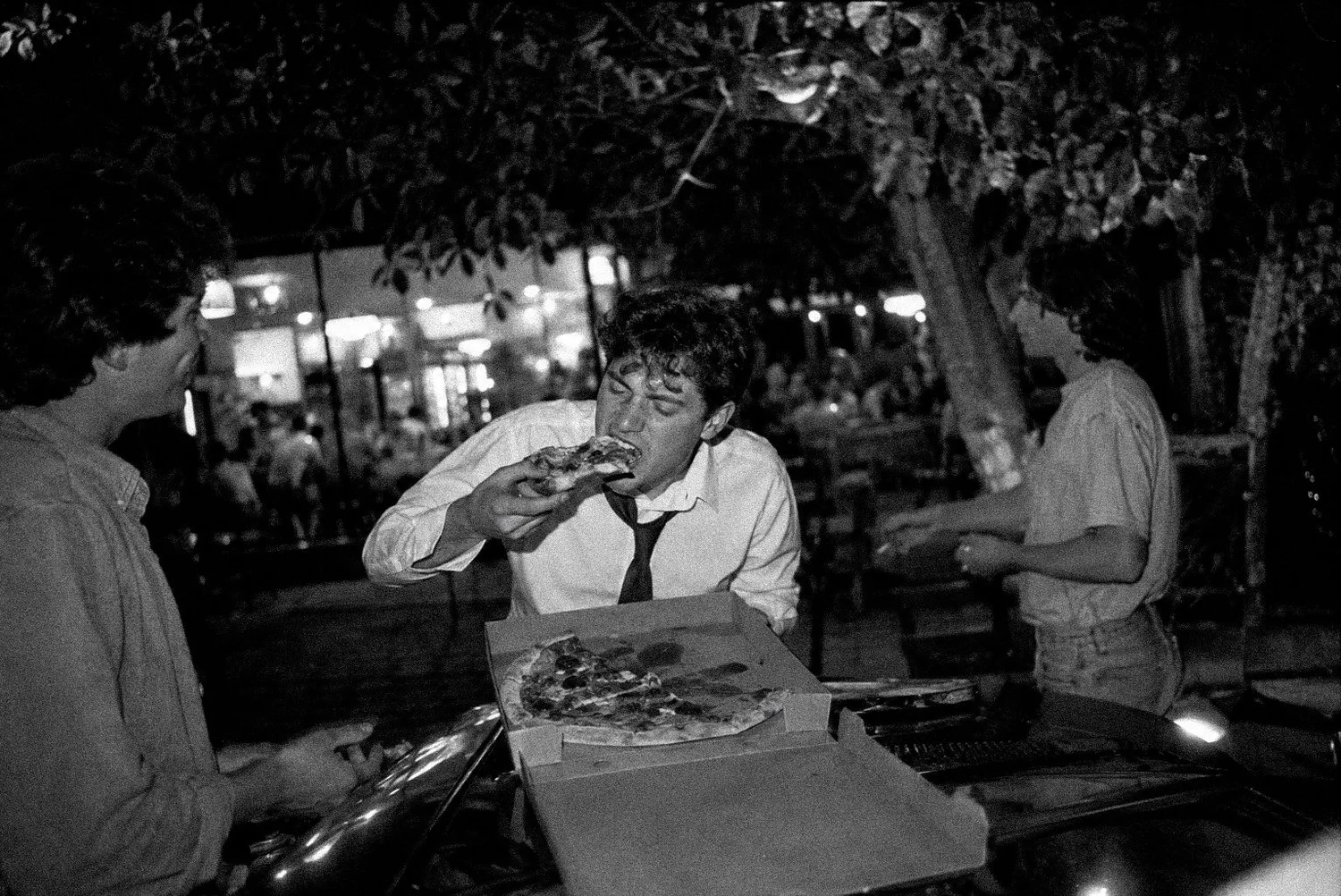 A young man in a shirt and tie eating a slice of pizza from an open box at an outdoor table at night.