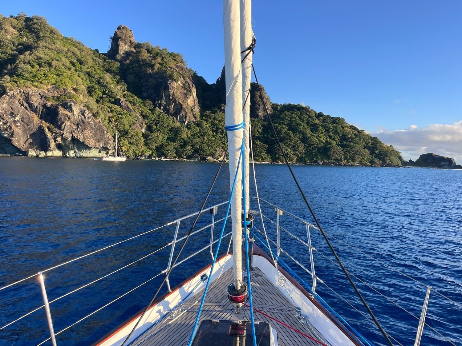 View over a yacht’s bow towards a rocky, tree-covered island across calm blue water
