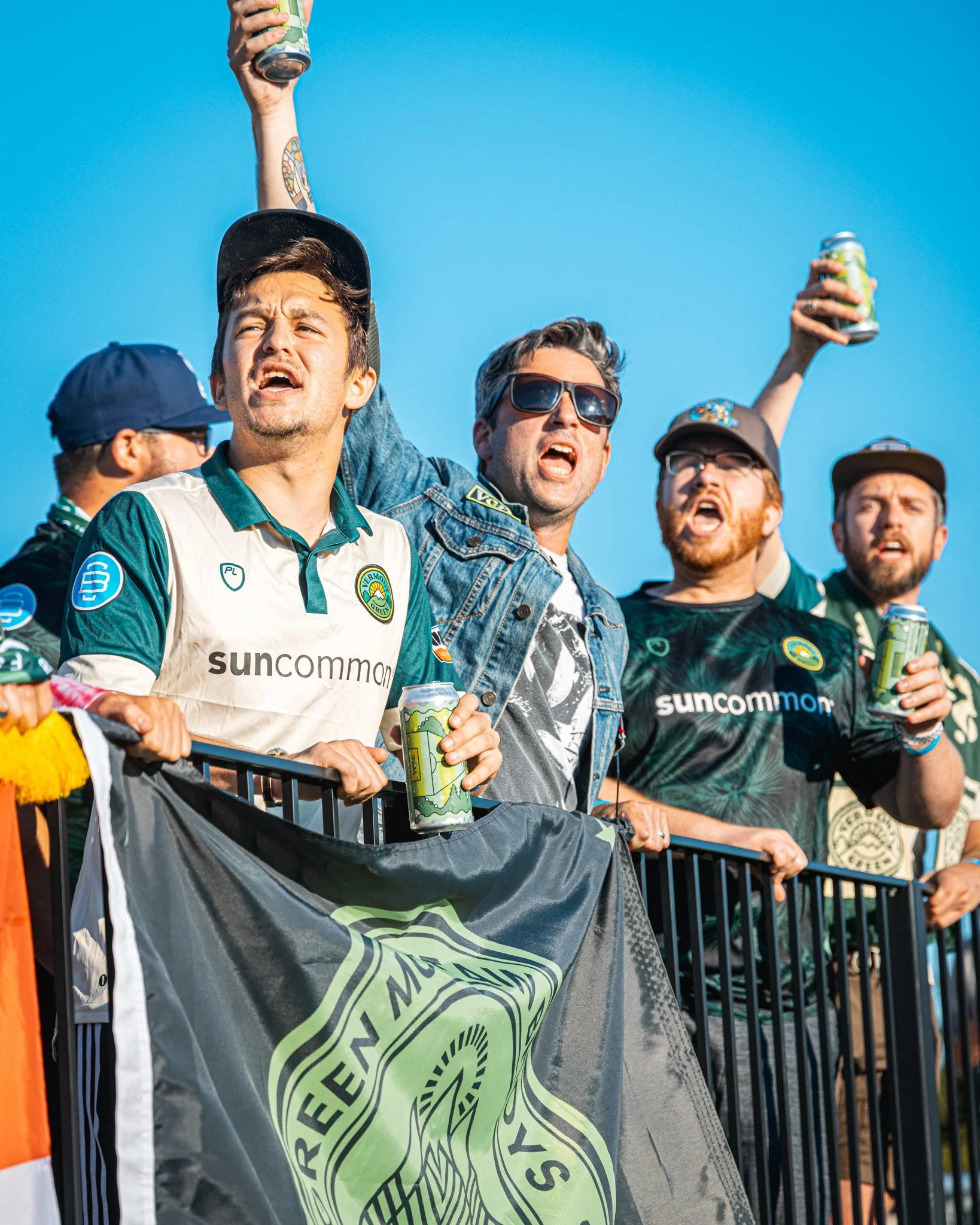 Vermont Green FC supporters cheering with Go Green Pale Ale cans, wearing jerseys with Burlington Beer Co. sleeve logos.