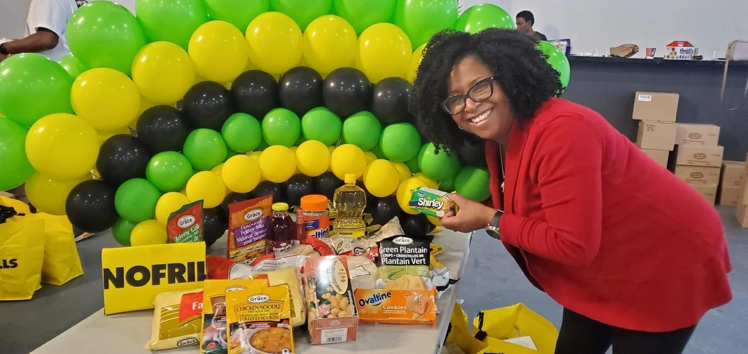 A woman smiling beside Afro-Caribbean Food Basket groceries under green, yellow and black balloons