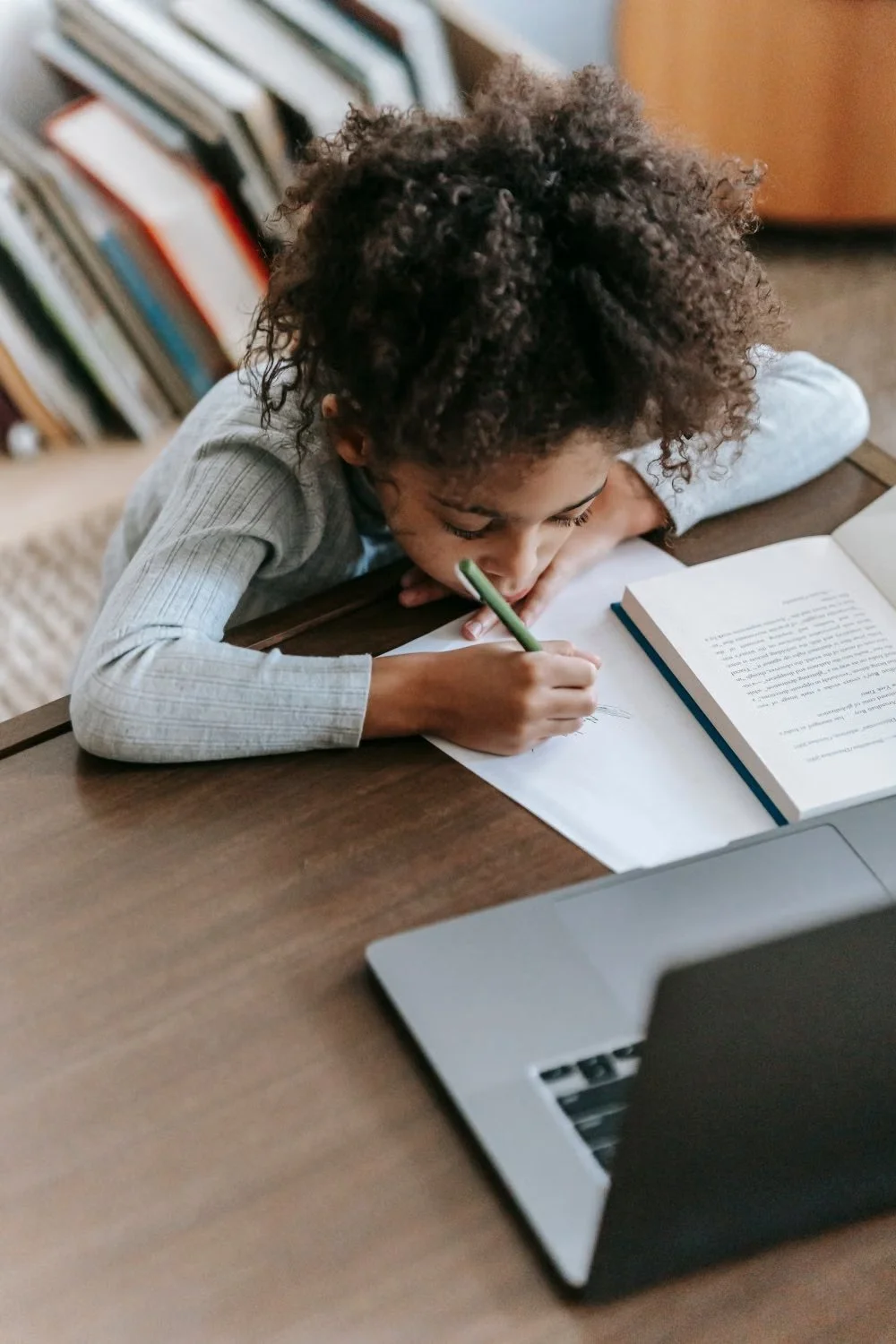 A child writing at a table with a book and laptop