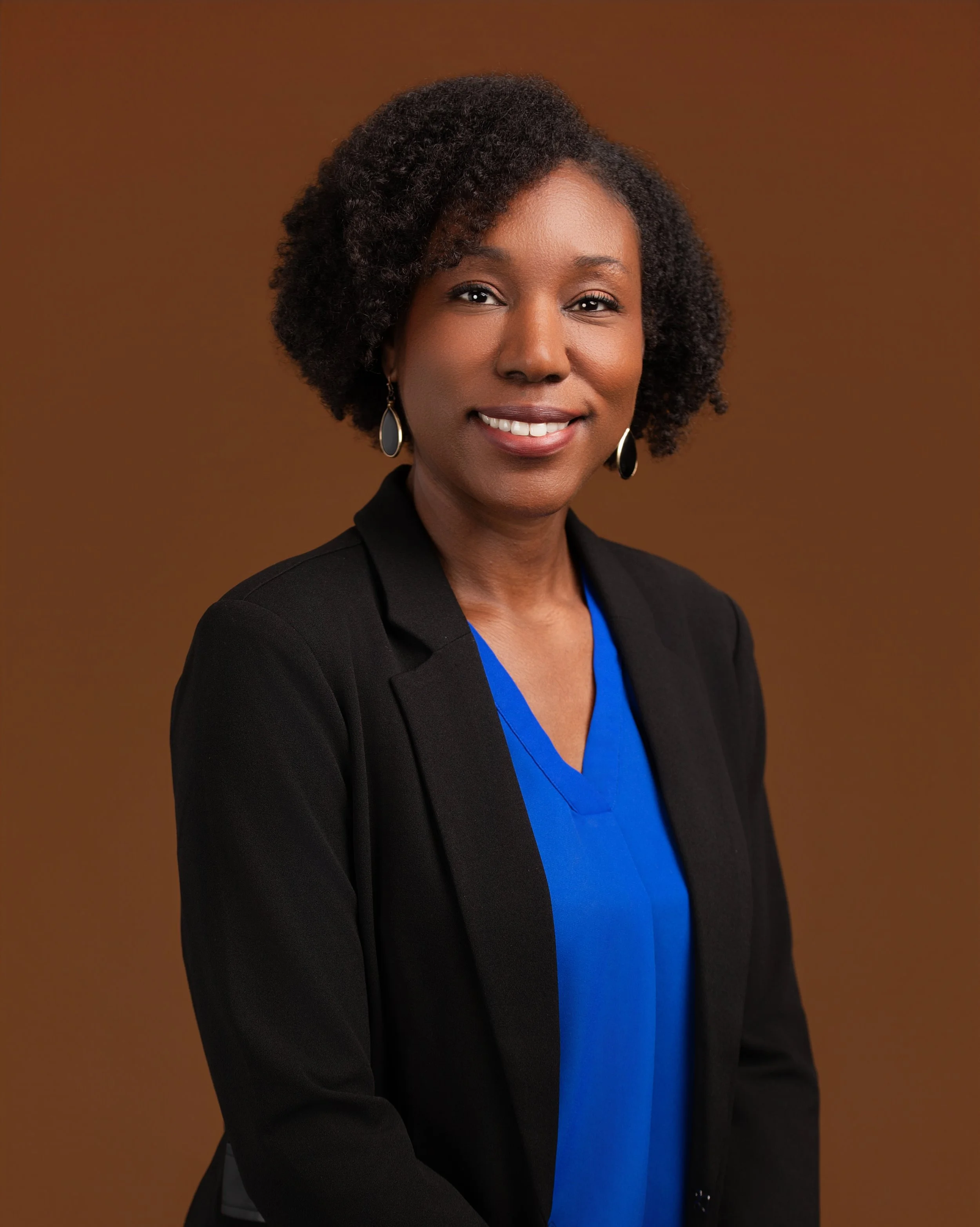 A headshot of a woman wearing a blue top and a black blazer in front of a brown backdrop.