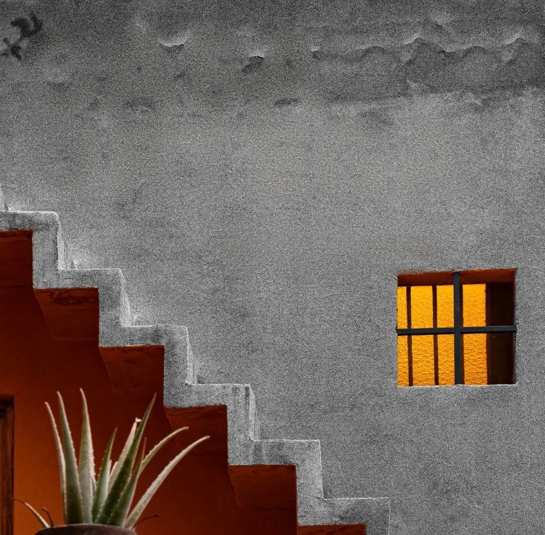 Escalera exterior sin barandal en un muro gris, con una ventana naranja