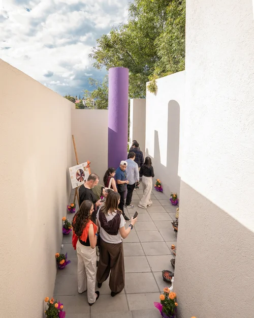 Ofrenda de 2025 en LEGORRETA: un pasillo de muros blancos con una columna morada y macetas de cempasúchil a los lados.