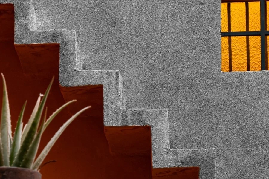 Jardines de Casa Ortega: escalera de concreto sin barandal junto a un muro gris, una ventana de vidrio amarillo y un agave en maceta.