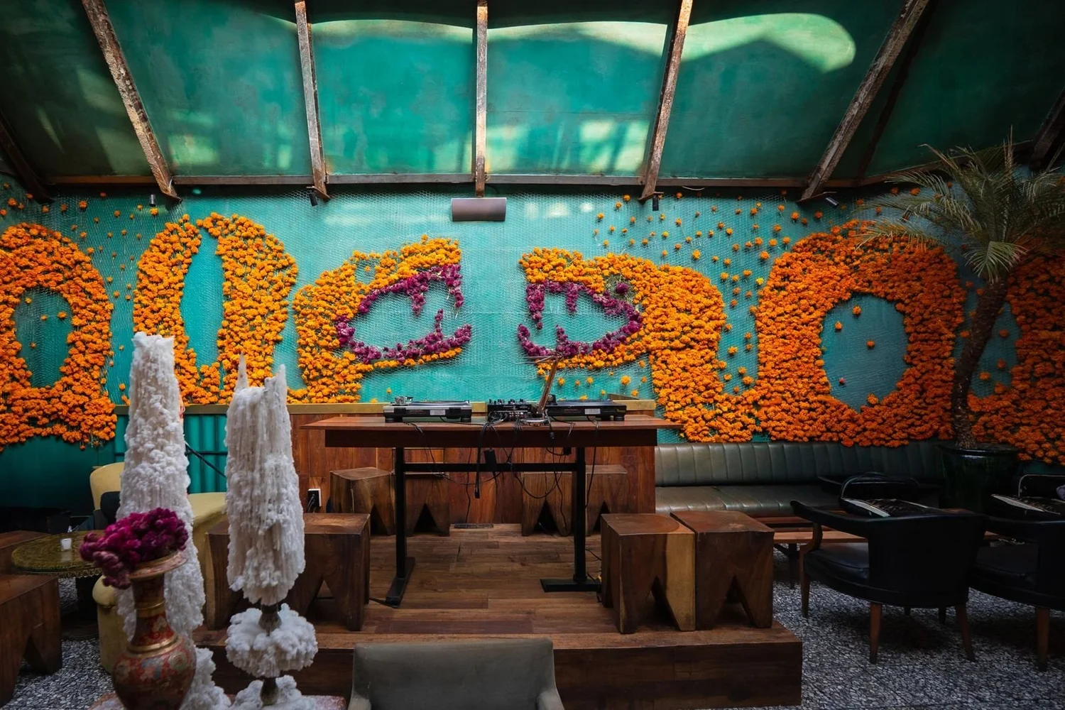 Muro turquesa cubierto de cempasúchil formando letras, con una mesa y dos piezas blancas al frente.
