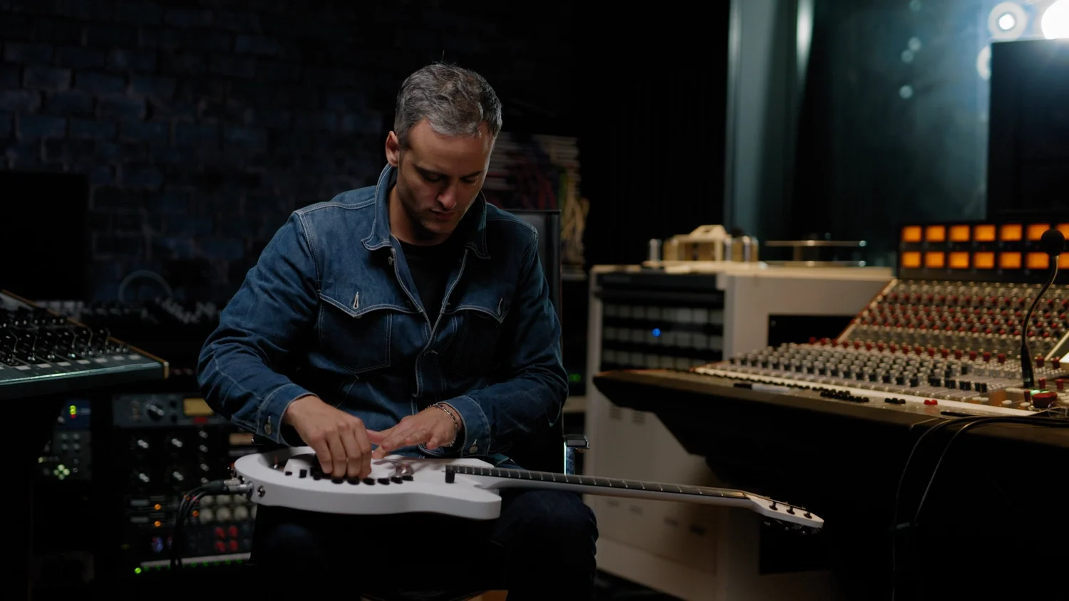 A musician seated in a recording studio with a white Circle Guitar across his lap, both hands on its controls, a mixing desk beside him