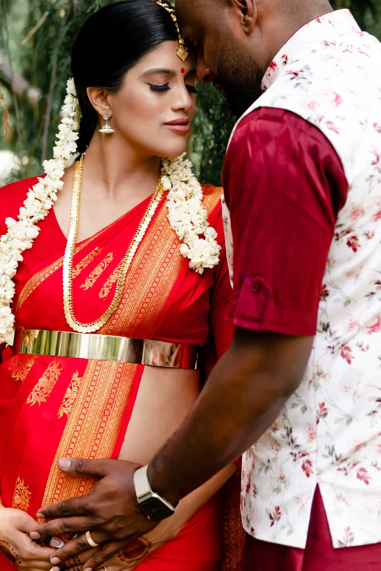An expectant couple in traditional dress rest their foreheads together, his hands over hers on her bump.